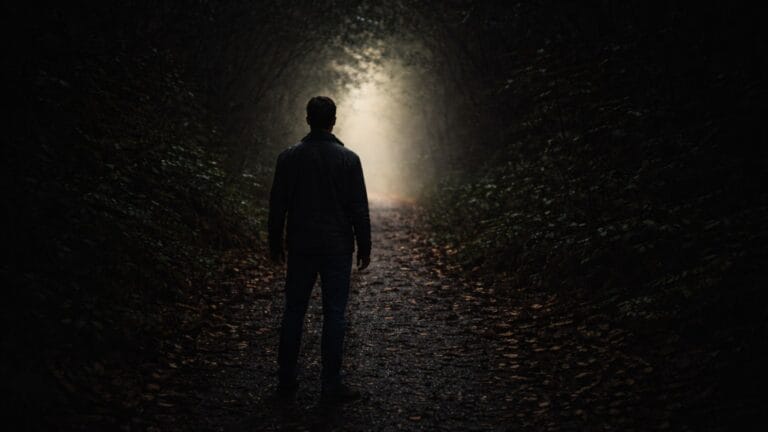A lone man stands on a dark forest path facing a distant light, visual for quotes about fear